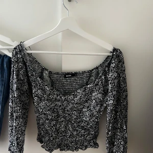 Blommig långärmad topp från Bik Bok - Supersöt svart och vit blommig topp från Bik Bok med lång ärm och smockad midja. Toppen har en lätt croppad passform, v-ringning och små volangdetaljer vid nederkanten. Perfekt för dig som gillar romantiska vibes och snygga mönster.