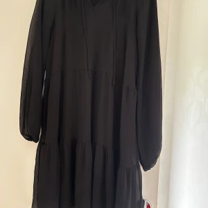 Svart volangklänning med snörning - En svart, luftig långklänning med långa ärmar och v-ringning. Klänningen har snörning vid halsen och flera volanglager som ger en avslappnad och trendig look. Perfekt för dig som gillar en enkel men snygg stil.