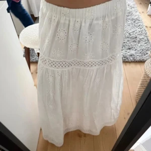 Vit broderad långkjol med spetsdetaljer - Superfin vit långkjol med broderade blommor och snygga spetsdetaljer. Kjolen har resår i midjan och är gjord i luftig bomull, perfekt för varma dagar. Den har en romantisk och bohemisk vibe med vackra hålbroderier och dekorativa band.