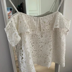 Vit spetsblus med volang - Superfin vit blus i spets med blommigt mönster och volangdetaljer vid axlarna. Blusen är kortärmad och har en luftig, romantisk känsla som passar perfekt till ljusa dagar. Snygg att styla med jeans eller kjol för en fräsch look.