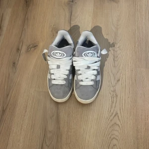 Adidas Campus 00s grå/vita sneakers - Säljer ett par Adidas Campus 00s sneakers i grå mocka med vita detaljer och klassiska tre ränder på sidorna. Skorna har breda vita skosnören, gummisula i brun ton och rund tå. Perfekta för dig som gillar retrostil och streetwear. Har bara använt de en gångs kan prata om priset.