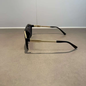 Svarta och guldiga Louis Vuitton solglasögon - Snygga solglasögon från Louis Vuitton med svart båge och guldiga detaljer längs ovansidan och skalmarna. Modellen har en klassisk pilotform med mörka glas och tydlig logga på sidan. Perfekt accessoar för att lyfta din stil till nästa nivå.