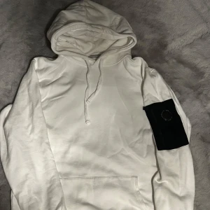 Vit hoodie från Snoot med ärmficka - Vit hoodie från Snoot i mjuk bomull med stor huva och snörning. På vänster ärm finns en svart ficka med dragkedja och Snoot-logga. Perfekt för dig som gillar stilrena plagg med coola detaljer.