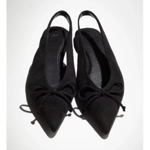 Svarta ballerinaskor med rosett H&M - Stilrena svarta ballerinaskor från H&M med spetsig tå och öppen häl. Skorna är i storlek 39 och aldrig använda, har prislapp kvar.