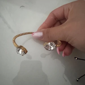 Carolina svedbom - Snyggt armband i guldtonad metall med tvinnad design och två stora, glittrande klara stenar i varje ände. Perfekt statement piece som drar blickarna till sig och ger en lyxig känsla till din outfit.