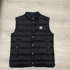 Moncler väst  - Svart dunväst med klassisk ståkrage och horisontella quiltade sömmar. Västen har tryckknappar framtill, två fickor med knappstängning och Monclers ikoniska logga på bröstet. Perfekt för lager-på-lager och snygg till både hoodie och t-shirt.