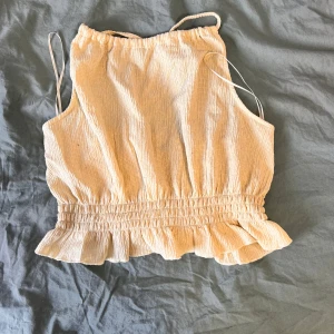 Gul topp med volangkant - Supersöt gul topp med smala axelband som korsas i ryggen. Toppen har en rynkad, elastisk midja och volangkant nedtill för en extra feminin touch. Perfekt för varma dagar och enkel att matcha med jeans eller kjol.