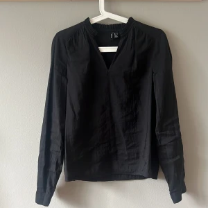Svart blus från Vero Moda, stl S - Snygg svart blus från Vero Moda med lång ärm och v-ringad hals. Blusen har en lätt glansig yta och resårdetalj vid halsringningen. Perfekt till jeans eller kjol för en stilren look.