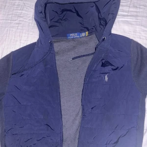 Blå hybrid jacka från Polo Ralph Lauren - Snygg mörkblå quiltad jacka från Polo Ralph Lauren med huva och dragkedja framtill. Jackan har snörning vid huvan och klassisk logga på bröstet. Ärmarna är i mjukare material och kroppen är lätt vadderad för extra värme. Perfekt för vår sommar höst, pris kan diskuteras