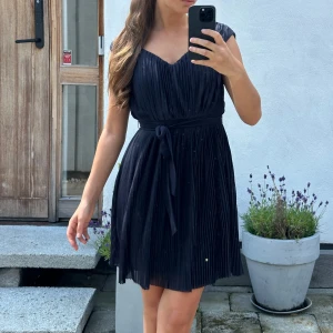 Mörkblå plisserad kortklänning med knyt - Supersnygg mörkblå plisserad klänning med v-ringning och knytband i midjan. Klänningen är kort, har cap-ärm och ett skirt, glansigt tyg som ger en lyxig känsla. Perfekt för dig som vill ha en stilren och trendig look.