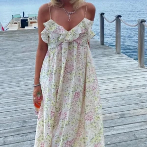 Blommig långklänning med volang - Romantisk långklänning med smala axelband och volang vid bysten. Klänningen är vit med blommigt mönster i rosa och grönt, har en luftig passform och är ärmlös. Perfekt för varma dagar och somriga vibes.
