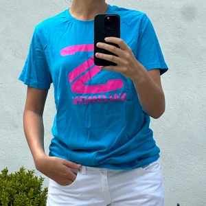 Blå Salomon t-shirt med rosa tryck - Snygg blå t-shirt från Salomon med stort rosa logotryck på bröstet. Klassisk rund hals och korta ärmar, perfekt för dig som gillar sportig stil. Skön och lätt i bomull, passar till jeans eller shorts för en chill look.
