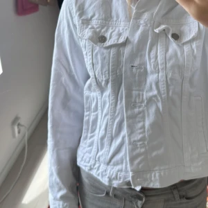 Vit jeansjacka med knappar - Snygg vit jeansjacka med klassisk passform och silverfärgade knappar framtill. Jackan har två bröstfickor med lock och knapp, samt markerade sömmar som ger en cool look. Perfekt att slänga över en hoodie eller t-shirt.
