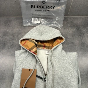 Burberry tröja  - Snygg ljusgrå hoodie från Burberry med klassiskt rutigt foder i huvan. Tröjan har dragkedja framtill, snörning i huvan och är tillverkad i mjuk bomull. Perfekt för dig som gillar stilrena och exklusiva plagg med en twist av ikoniskt mönster.