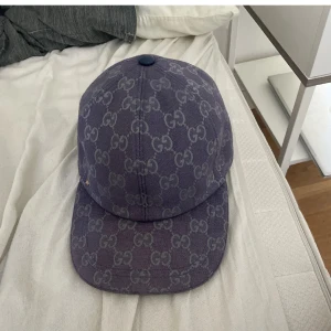 Lila Gucci keps med GG-mönster - Snygg lila keps från Gucci med klassiskt GG-mönster över hela kepsen. Justerbar rem baktill och böjd skärm. Perfekt för dig som vill sticka ut med en exklusiv accessoar från ett av världens mest ikoniska modehus.