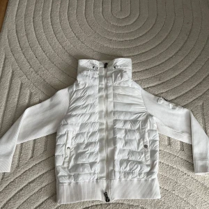 Vit pufferjacka från Moncler - Snygg vit pufferjacka från Moncler med ribbstickade ärmar och quiltad front. Jackan har hög krage, huva och dragkedja framtill. Fickor med tryckknappar och klassisk Moncler-logga på ärmen. Perfekt för dig som vill ha en clean och sportig look.