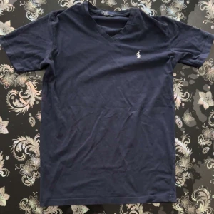 Mörkblå Polo Ralph Lauren T-shirt - Snygg mörkblå t-shirt från Polo Ralph Lauren med klassisk v-ringning och vit broderad logga på bröstet. Tillverkad i mjuk bomull och har korta ärmar. Perfekt för en clean och stilren look.