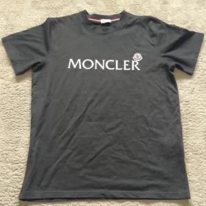 Svart Moncler t-shirt med logga - Svart t-shirt från Moncler med klassisk rund hals och vit Moncler-logga tryckt på bröstet. T-shirten har korta ärmar och en liten broderad Moncler-patch på vänster sida. Tillverkad i mjuk bomull för en skön känsla.