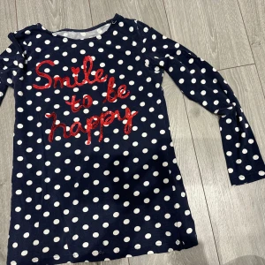 Mörkblå prickig klänning med text för barn  - Mörkblå kortklänning med vita prickar och röd paljetttexten 'Smile to be happy' framtill. Långärmad och i mjukt material, perfekt för dig som gillar lekfull stil och coola detaljer. Skulle nog passa 4-6 åringar