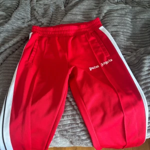 Röda Palm Angels mjukisbyxor L - Palm Angels röda mjukisbyxor med vita sidoränder och logga på låret. Byxorna har elastisk midja och raka ben. Tillverkade i 100% polyester för en sportig vibe. Perfekta för dig som gillar streetwear och vill sticka ut.