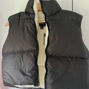 Svart dunväst från Boohoo - Svart dunväst från Boohoo med hög krage och vit teddyfoder på insidan. Västen har en dragkedja framtill och är quiltad för extra volym. Perfekt att slänga över en hoodie för en stilren look när det är kallt ute.