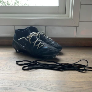 Nike Phantom Luna svarta fotbollsskor PRO. svarta original snören ingår  - Svarta Nike Phantom Luna fotbollsskor med högt skaft i stickad syntet och vita snören. Skorna har rund tå, platt sula med dobbar och diskret blå Nike-logga på sidan. Perfekta för dig som vill ha stilrena och tekniska skor på planen. Svarta Original snören ingår 