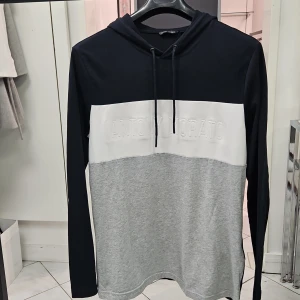 Blockfärgad hoodie Antony Morato - Snygg hoodie från Antony Morato i storlek L. Tröjan har blockfärger i svart, vitt och grått med broderad logga framtill. Långärmad modell med huva och snörning. Perfekt för en chill och stilren look. Hoodie är en aning mindre i storleken. Material: Bomull 