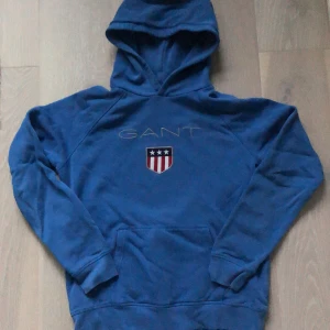 Blå hoodie från GANT med brodyr - Snygg blå hoodie från GANT med broderad logga och sköld på bröstet. Tröjan har huva, magficka och långa ärmar. Perfekt för dig som gillar klassisk och sportig stil. Mjuk och skön i bomullsmix. Tröjan är endast använd ett fåtal gånger och säljes nu eftersom den inte längre passar mig. Priset går att diskuteras.
