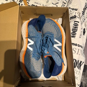 Blå New Balance sneakers med orange detalj - Snygga blå sneakers från New Balance med vita loggor på sidorna och orange detaljer runt sulan. Ovandelen är i mesh för bra andningsförmåga och sulan är tjock och bekväm. Perfekta för dig som gillar sportig stil och vill ha något som sticker ut.