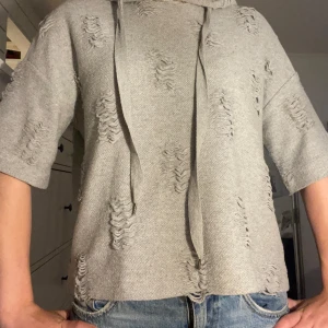 Skitsnygg Grå hoodie med slitna detaljer - Grå hoodie med korta ärmar och coola slitna detaljer över hela tröjan. Tröjan har huva och snörning framtill, loose fit och är tillverkad i ett mjukt stickat material.Okänt varumärke då den är köpt på vi Vinted!💕