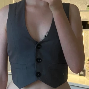 Mörkgrå väst från Gina Tricot - Snygg mörkgrå väst från Gina Tricot med djup v-ringning och tre knappar framtill. Västen har två dekorativa fickor och är figursydd för en modern look. Perfekt att styla över en skjorta eller t-shirt för en trendig vibe.