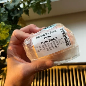 Shake Ya Bum Bath Bomb - En rund badbomb i orange, rosa och vit färg från Shake Ya Bum. Kommer inslagen i genomskinligt papper med etikett. Perfekt för ett bubbligt och färgglatt bad, doftar fräscht och ger en lyxig känsla i badkaret.
