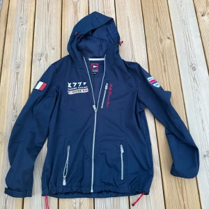 Marinblå vindjacka från Marine Sailing - Snygg marinblå vindjacka med huva från Marine Sailing. Jackan har broderade detaljer, dragkedjor framtill och på fickorna samt coola patches på ärmarna. Perfekt för dig som gillar en sportig och nautisk stil. Justerbar nederkant med snörning och tryck på ryggen.