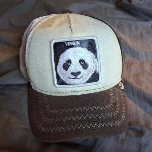 Beige och brun keps The Farm Panda - Snygg keps från The Farm med beige front, brun skärm och vit/svart mesh baktill. Framtill sitter ett broderat panda-märke med texten 'VIRGIN'. Skärmen har dekorativa zigzag-sömmar. Perfekt för dig som vill sticka ut med en unik accessoar.
