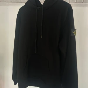 Svart hoodie från Stone Island - Svart hoodie från Stone Island med klassisk patch på vänster ärm. Tröjan har huva med snörning och en stor magficka framtill. Perfekt för dig som gillar streetwear och vill ha en stilren look.