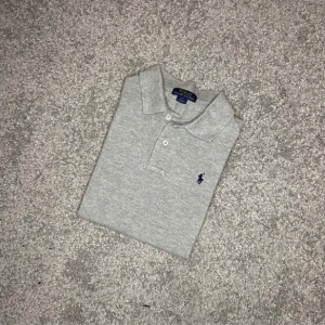Ralph lauren långärmad piké  - Stilren grå långärmad piké från Ralph Lauren – en perfekt kombination av sportig elegans och komfort. Tillverkad i mjuk bomull med klassisk krage, knappar och den ikoniska broderade loggan på bröstet. Ett mångsidigt plagg som passar året runt – lika snygg till jeans som chinos. Inga fläckar eller slitage – fräsch och redo för användning!