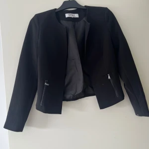 Svart kavajjacka från ONLY, stl 34 - Stilren svart kavajjacka från ONLY med lång ärm och snygga dragkedjor vid fickorna. Jackan har en rak, lite kortare passform och är fodrad för extra komfort. Perfekt för dig som gillar en clean look med edge. Kontakta för övriga frågor.