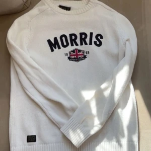 Vit stickad tröja från Morris - Vit stickad tröja från Morris med broderad logga och brittisk flagga framtill. Tröjan har rund hals, långa ärmar och ribbade muddar vid ärmslut och nederkant. Perfekt för dig som gillar klassisk och stilren look.