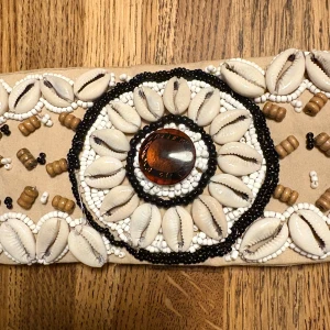 Mockabälte med snäckor och pärlor - Unikt beige bälte dekorerat med vita snäckor, bruna  och svarta samt vita småpärlor. Bältet har ett stort runt mönster i mitten med en brun glansig sten och knyts med en flätad snodd med tofsar. Perfekt för att ge din outfit en boho-vibe.