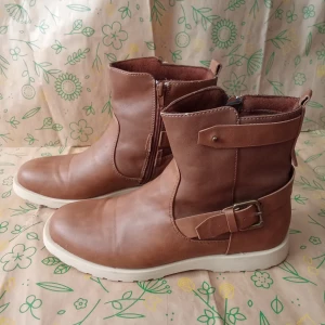 Bruna boots med spänne och dragkedja, 38 - Snygga bruna boots i skinnimitation med vit sula, rund tå och dekorativt spänne på sidan. Dragkedja på insidan för enkel påtagning. Perfekta för höst och vinter, passar till både jeans och kjol. Bekväm platt sula och stilren design. Kan skickas med extra pris via post.