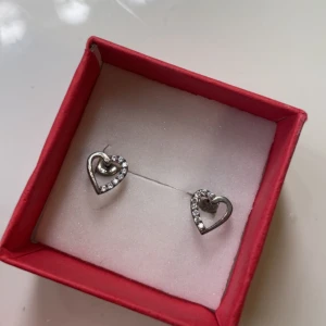 Hjärtformade örhängen i silver - Snygga örhängen- som inte rostar i form av hjärtan i silverfärg med glittriga stenar längs ena sidan. Köpte från sidan chanti för väldigt längesedan och varan säljs inte längre. Kommer i en röd ask. Orginalpris: 1050kr