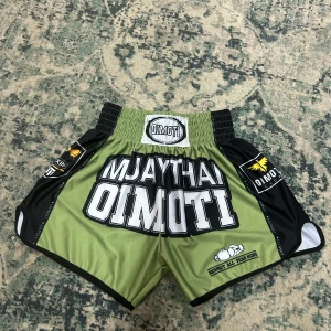 Muay Thai shorts från Oimoti - Grymma Muay Thai shorts från Oimoti i olivgrönt och svart med stora vita tryck och coola detaljer. Bred resår i midjan för extra komfort och rörelsefrihet. Perfekta för träning eller match, med texten 'RESPECT ALL FEAR NONE' och snygga loggor på sidorna.