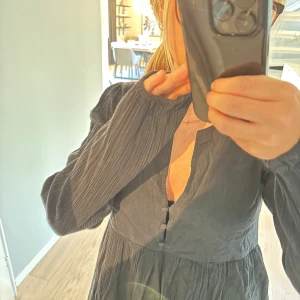 Svart kort klänning med knappar - Supersöt svart klänning med lång ärm och knappar framtill. Den har en lätt v-ringning, flowig kjol och är tillverkad i ett tunt, skönt tyg som ger en avslappnad vibe. Perfekt för dig som gillar en enkel men stilren look.