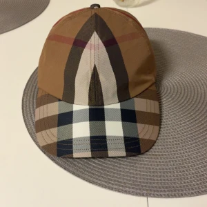 Burberry rutig keps  - Snygg keps från Burberry i klassiskt rutigt mönster med bruna, svarta, vita och beige toner. Broderad Burberry-logga baktill och justerbart spänne i metall. Tillverkad i 100% bomull för en skön och lyxig känsla.