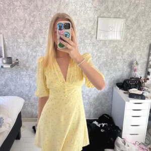 Gul blommig kortklänning från Adoore - Superfin gul kortklänning från Adoore med små blommor i grönt och vitt. Klänningen har v-ringning, knappar framtill, puffiga trekvartsärmar och lätt utsvängd kjol. Perfekt för sommarens alla tillfällen! Helt oanvänd med prislapp 