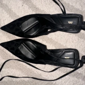 Svarta leopardmönstrade pumps från Zara - Snygga svarta pumps från Zara med spetsig tå och leopardmönster i sammet. Skorna har hög klack, knytband runt vristen och en elegant, modern design. Perfekta för dig som vill sticka ut med en trendig och edgy look.