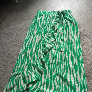 Grön zebramönstrad omlottkjol från Vila - Säljer en lång omlottkjol från Vila i grönvitt zebramönster. Kjolen har volang längs kanten och knyts i midjan med en rosett. Perfekt för dig som vill sticka ut med en färgglad och trendig look.
