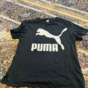 Svart Puma t-shirt med stor logga - Svart t-shirt från Puma i storlek L med stor vit logga och puma-tryck på bröstet. Klassisk rund hals och korta ärmar. Tillverkad i mjuk bomull, perfekt för en sportig och avslappnad look.