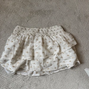 Vit volangkjol med guldprickar från Zara - Supersöt kort kjol från Zara i vitt med glittriga guldprickar och volanglager. Resår i midjan för skön passform. Perfekt för dig som vill ha en lekfull och trendig look med extra detaljer. Lågmidjade 