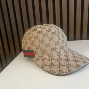 Gucci keps med GG-mönster - Pris kan diskuteras. Snygg keps från Gucci i beige och brunt med klassiskt GG-monogram över hela kepsen. På sidan finns den ikoniska gröna och röda randen. Kepsen är tillverkad i bomull och har en böjd skärm. Perfekt för dig som vill sticka ut med en lyxig accessoar.
